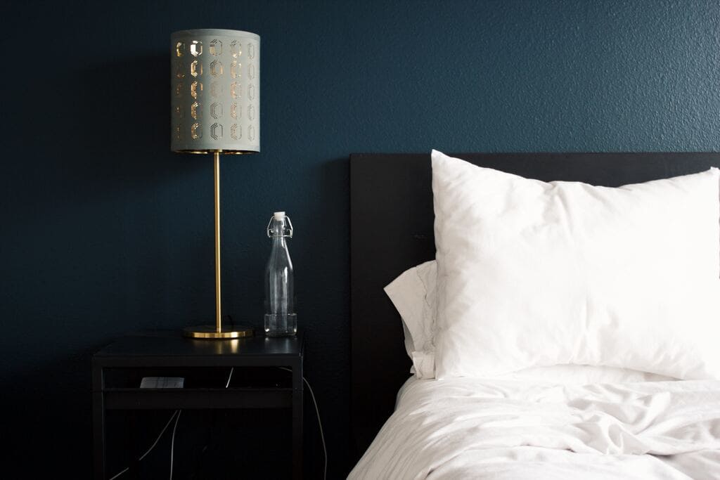 Dormitorio de hotel con cama negra y sabanas blancas. Mesa de noche con lampara moderna y botella de cristal. Fondo azul.